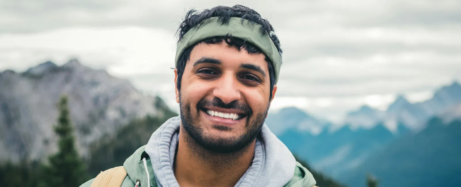 Smiling man wearing green hoodie and headband with backpack in mountainous outdoor setting under cloudy sky