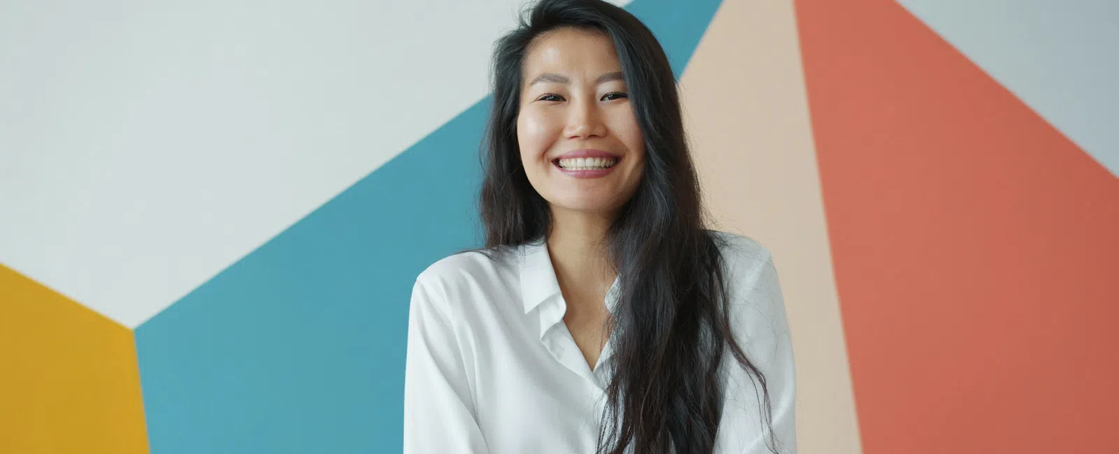 Smiling Asian woman with long black hair wearing a white shirt standing against a colorful geometric wall.