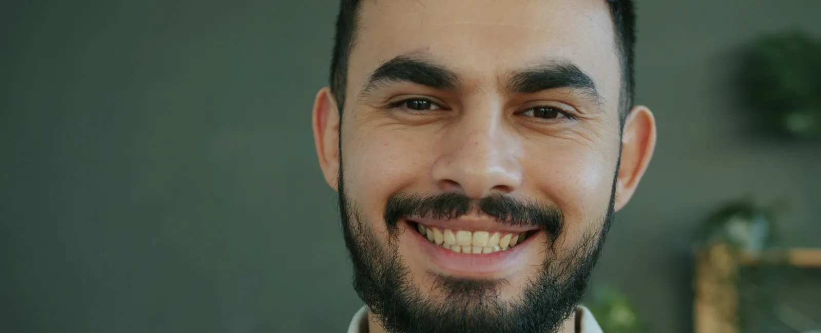 Smiling man with beard and mustache wearing a light gray shirt against a blurred indoor background
