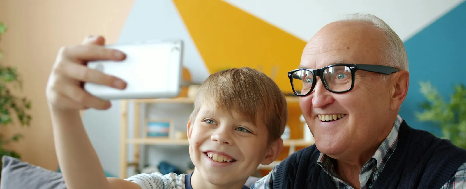 Young boy and elderly man smiling together while taking a selfie indoors with colorful geometric wall background.