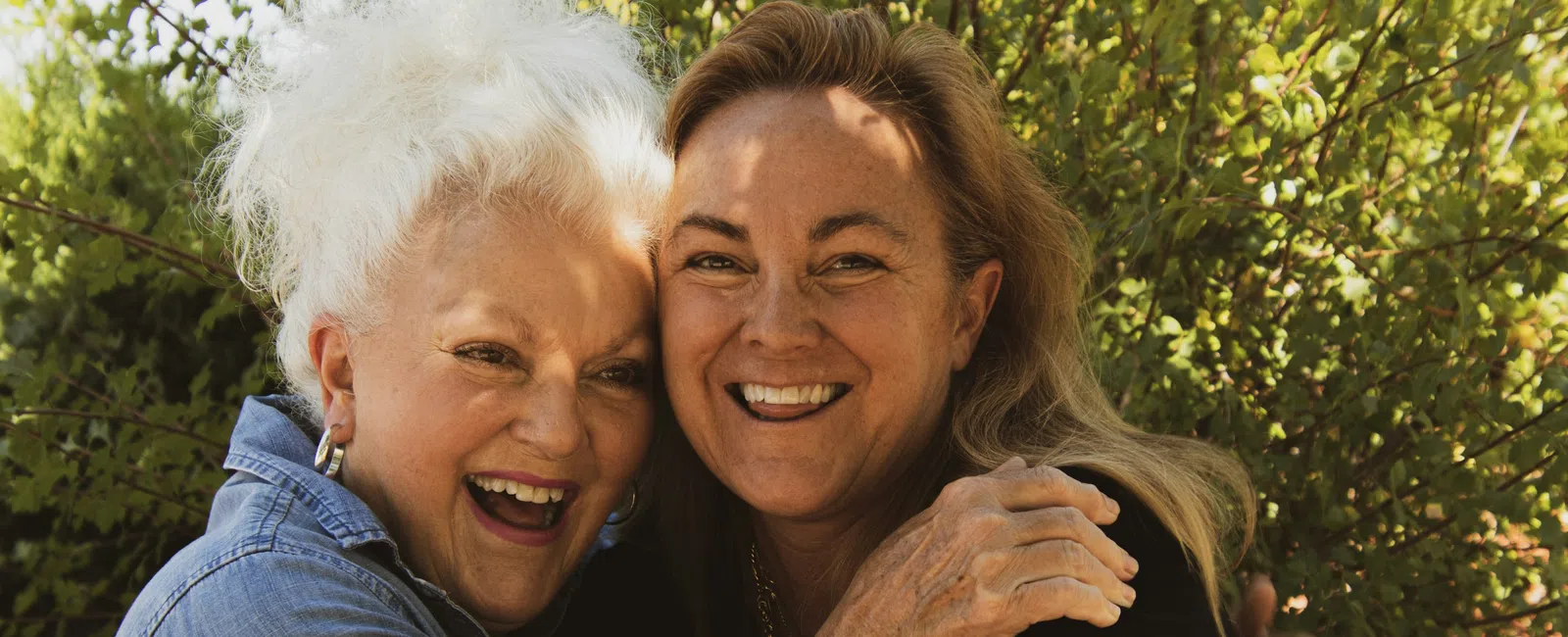 Two smiling women, one elderly with white hair and one middle-aged, embracing in a garden outdoors.