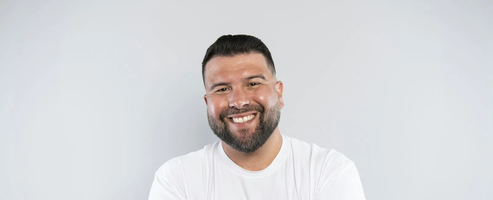 Smiling man with beard and short hair wearing white t-shirt standing with arms crossed on light gray background