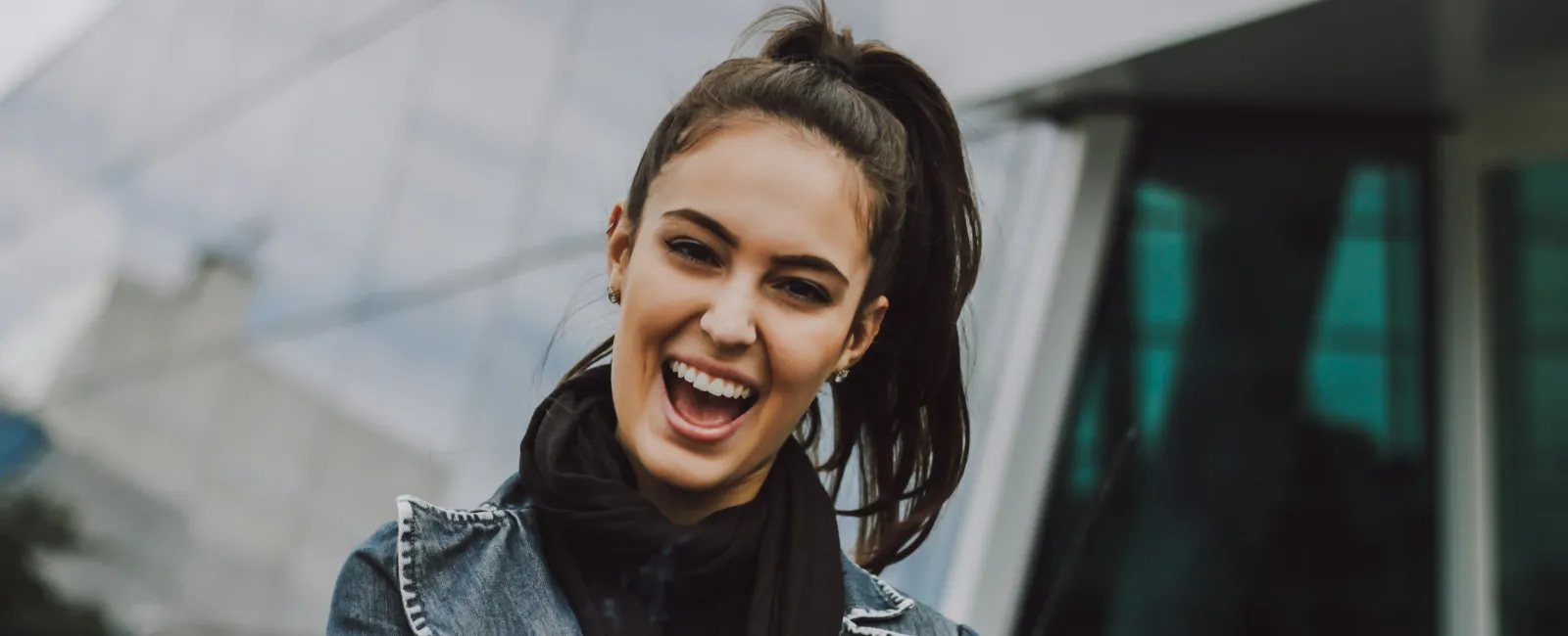 Smiling woman in denim jacket and scarf, standing outdoors with modern architecture in the background.