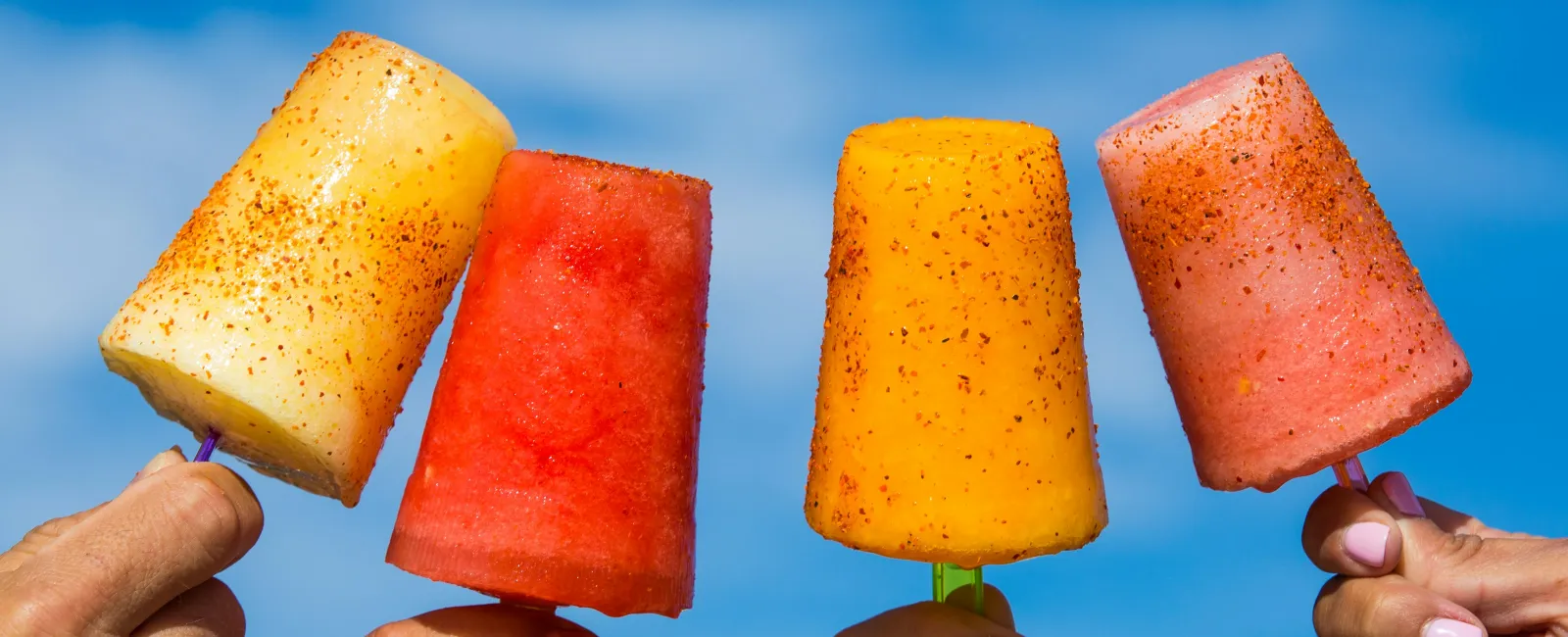 Four colorful fruit popsicles held up against a clear blue sky, sprinkled with chili powder.