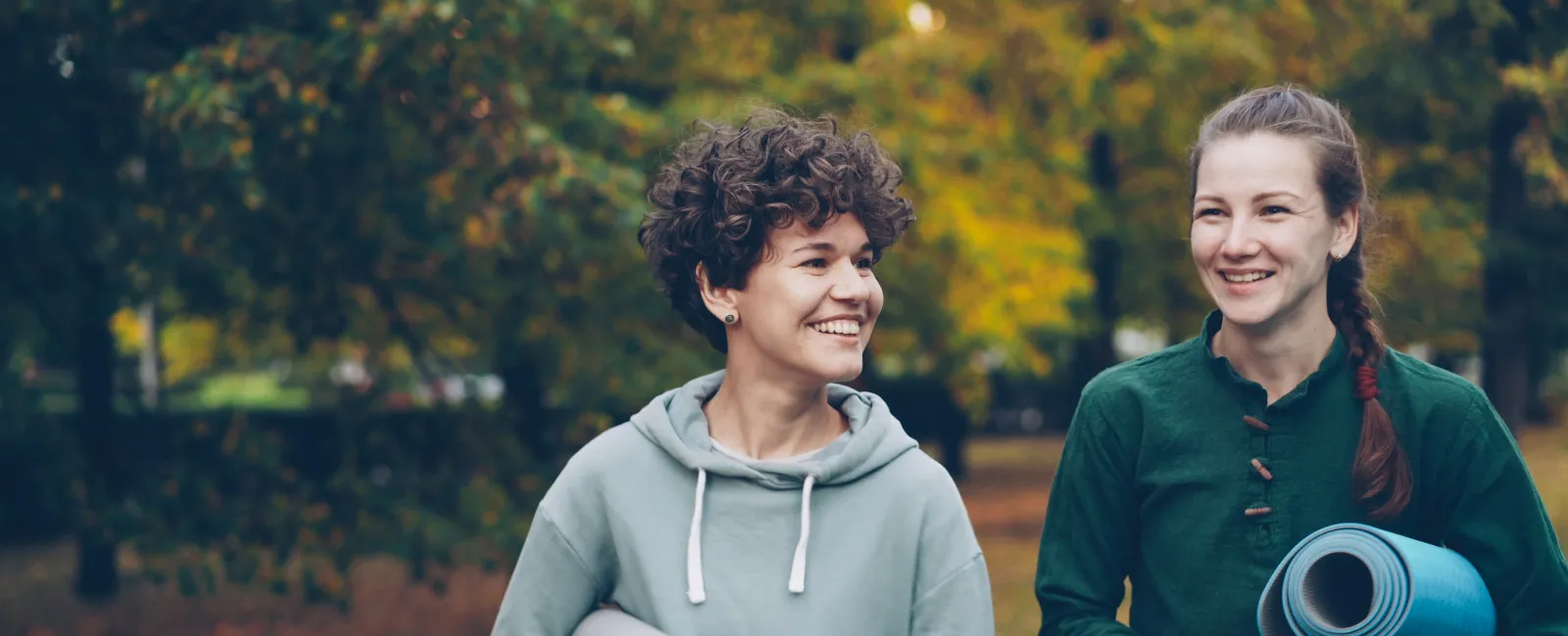 Two women smiling and holding yoga mats walking outdoors in autumn with fallen leaves and trees.