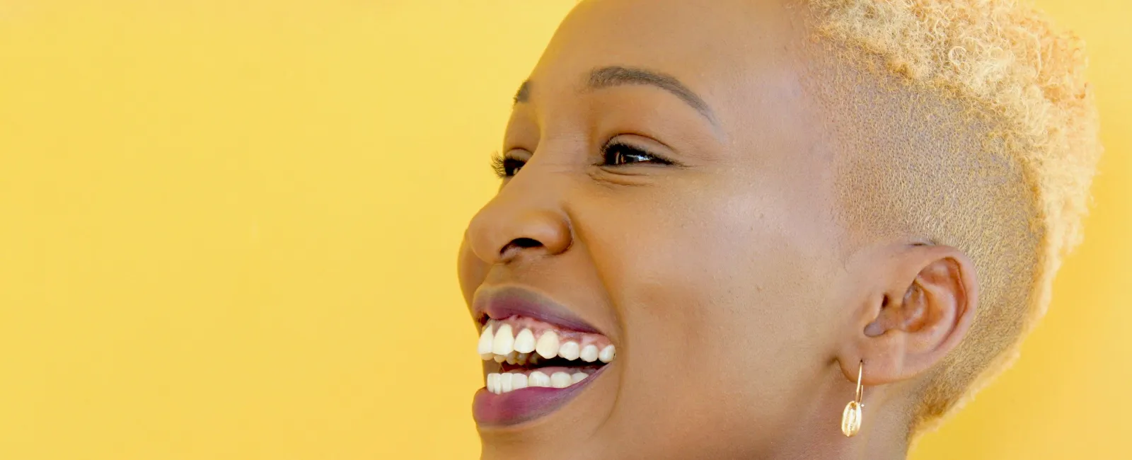 Close-up side view of a smiling woman with short blonde hair and gold earrings against a yellow background