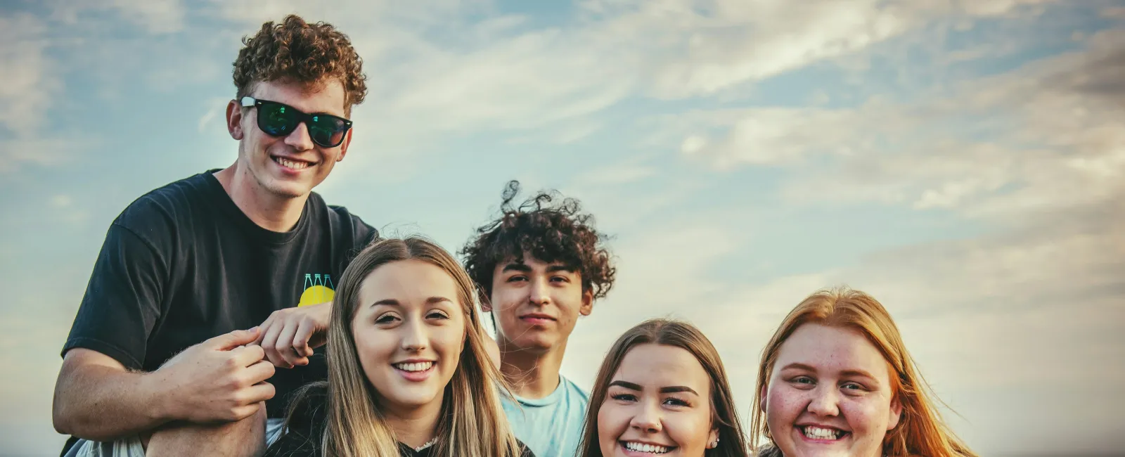 Group of five smiling young friends posing outdoors by the ocean during sunset golden hour.