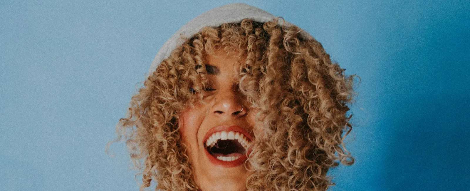 Laughing woman with curly hair wearing a gray hoodie against a blue background