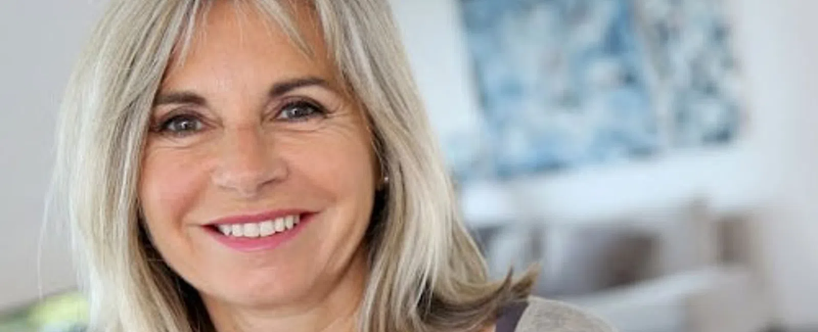 Smiling middle-aged woman with blonde hair in a casual top in a bright, modern living room setting.