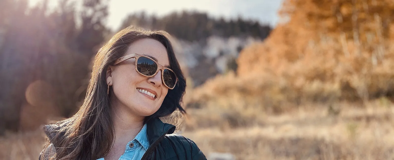 Smiling woman wearing sunglasses and jacket enjoying sunny autumn nature outdoors with trees in background