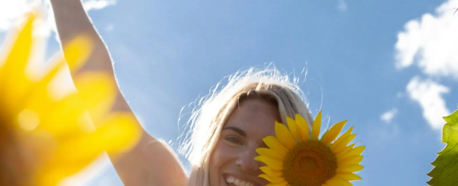 Smiling woman holding sunflower in a sunny field with blue sky and clouds, wearing striped dress.