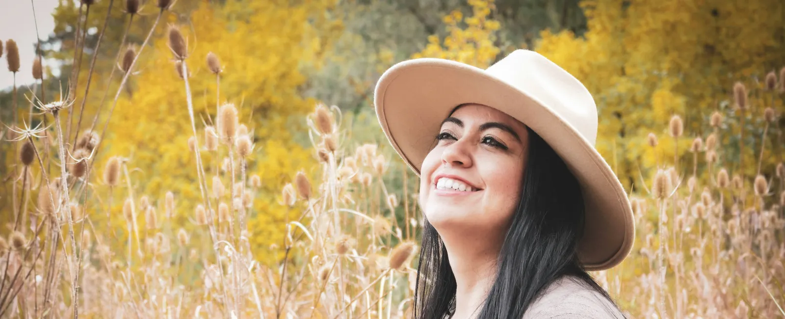 Smiling woman wearing a hat and cardigan sitting in a field with autumn foliage in the background
