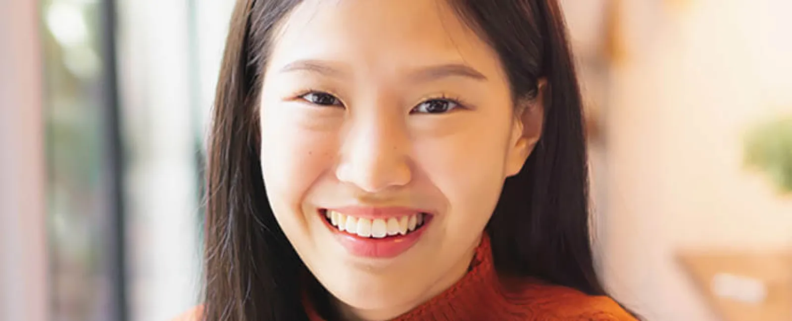 Smiling young woman wearing an orange sweater in a cozy, well-lit indoor setting.