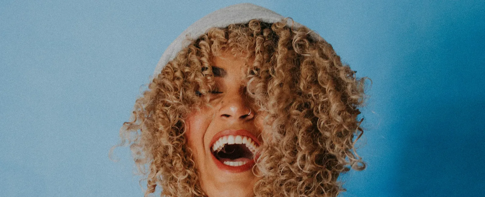 Young woman with curly hair laughing and holding hoodie strings against a blue background.
