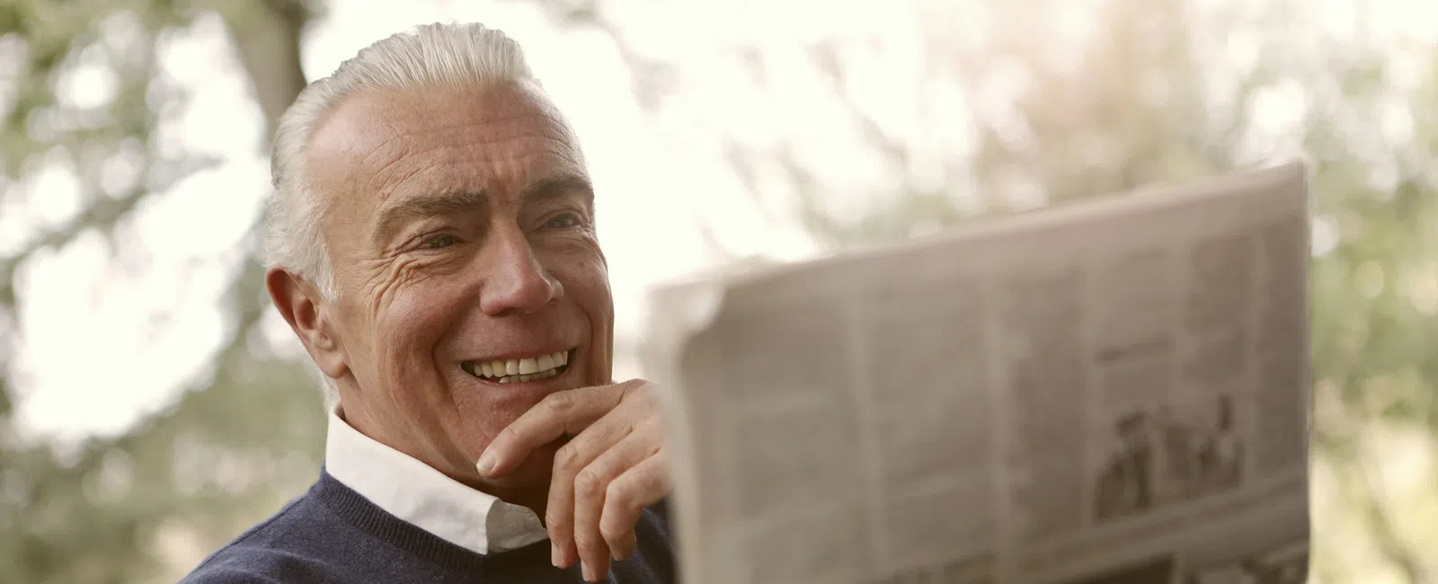 Smiling elderly man reading a newspaper outdoors with trees in the background in natural light.