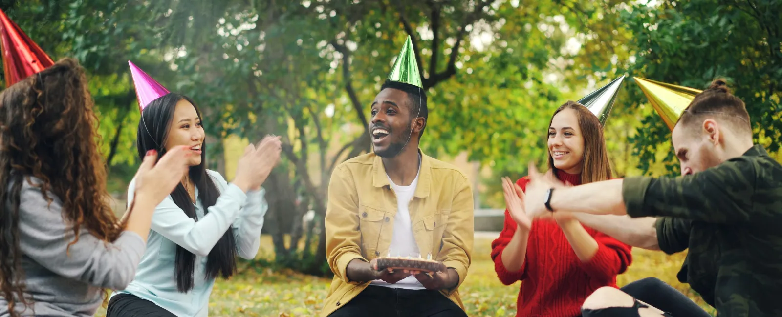 Group of friends wearing party hats celebrating a birthday at an outdoor picnic with cake and snacks