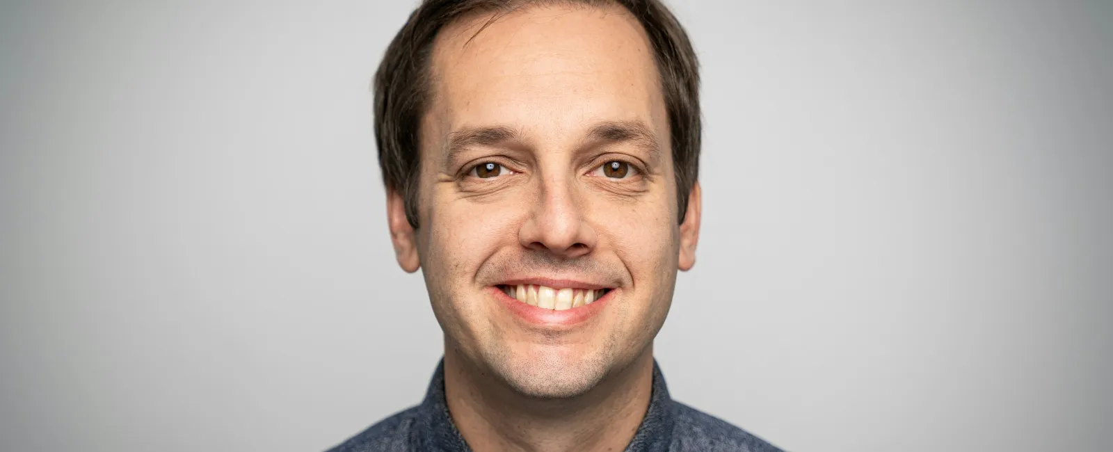 Smiling man with short brown hair wearing a blue button-up shirt against a light gray background