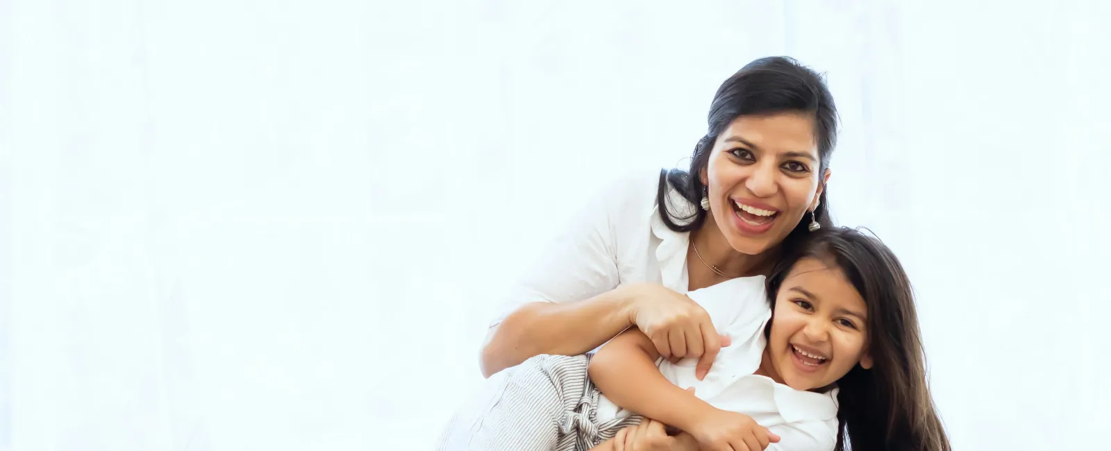 Mother and daughter smiling and hugging each other while sitting on a gray couch in bright room.