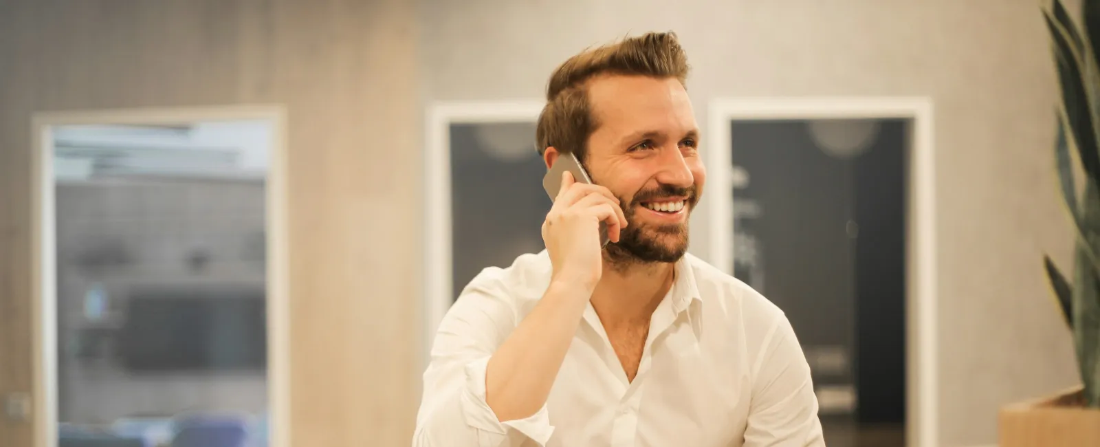 Smiling man in white shirt using phone and laptop at wooden desk with coffee cup in modern office space