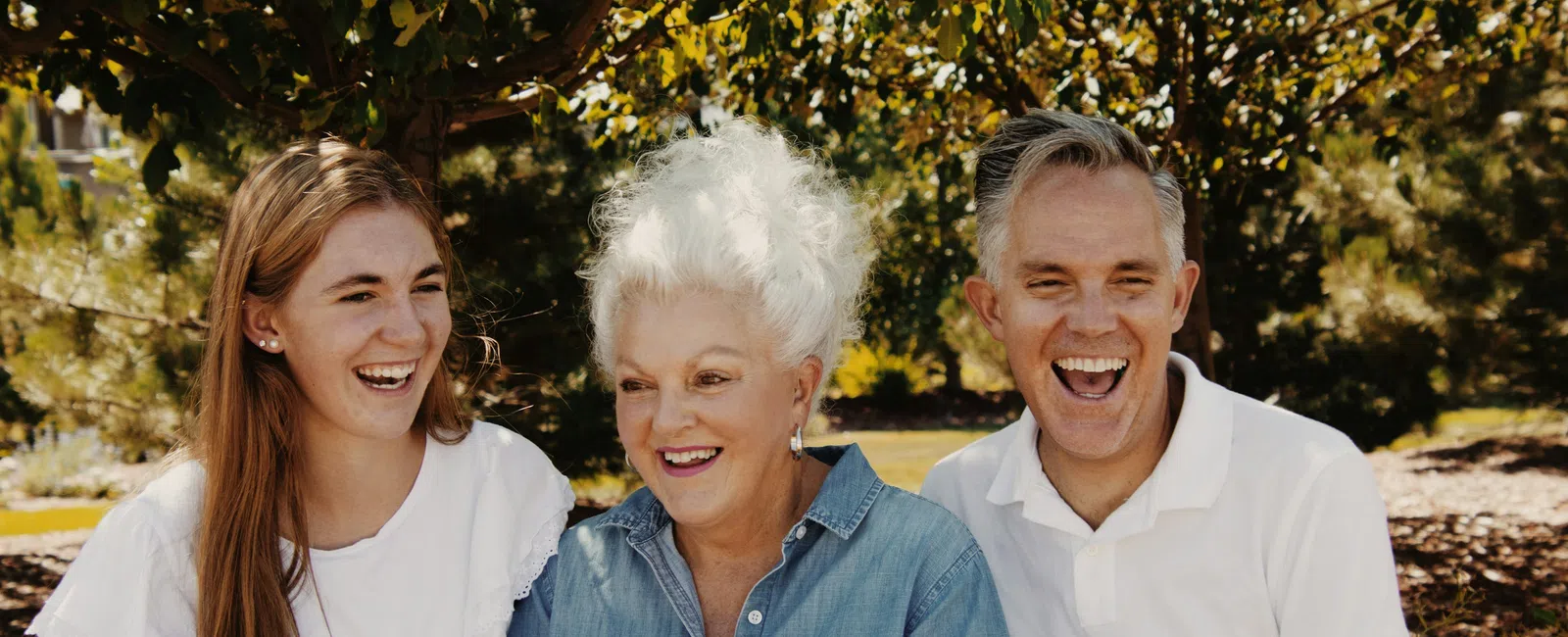 Three generations of a family smiling and laughing together outdoors under a tree on a sunny day.