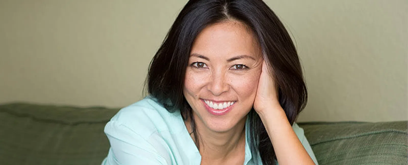 Smiling woman with dark hair in light blue blouse relaxing on green couch in cozy living room.