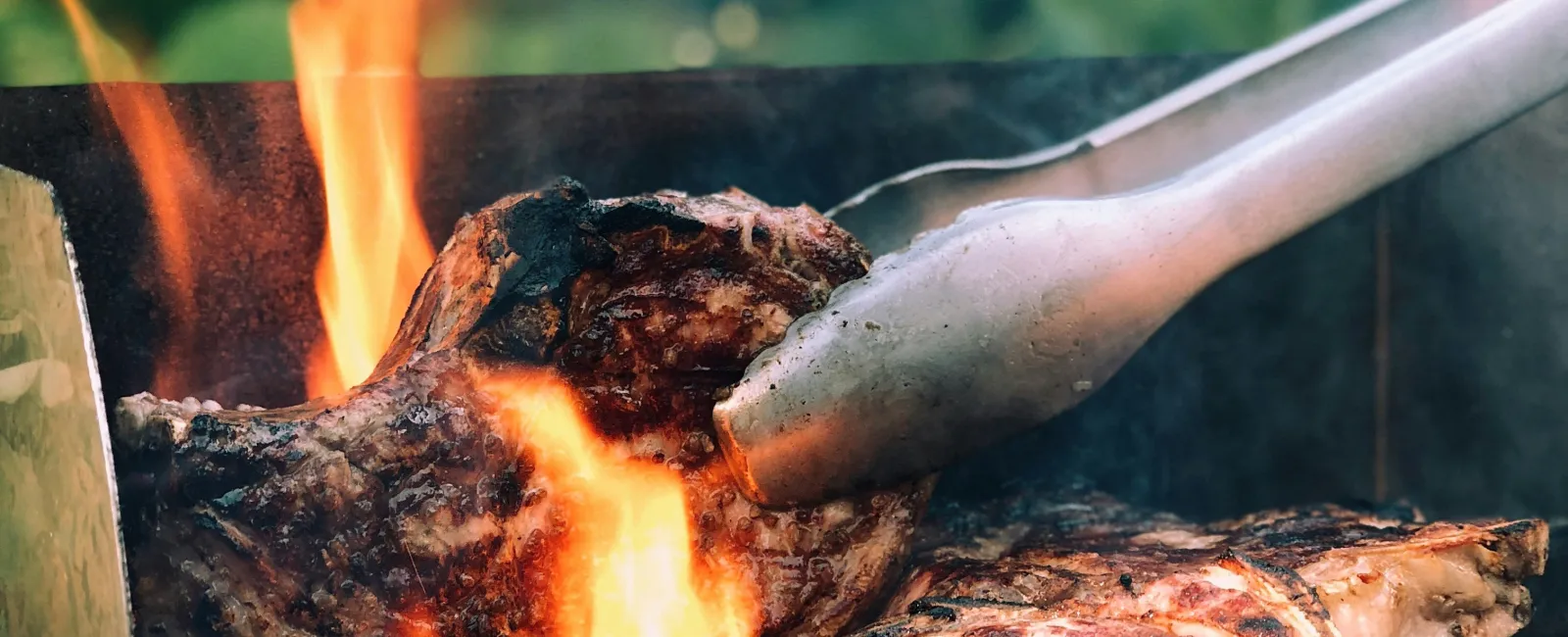 Close-up of thick steaks grilling over hot flames on a charcoal barbecue with tongs flipping the meat.