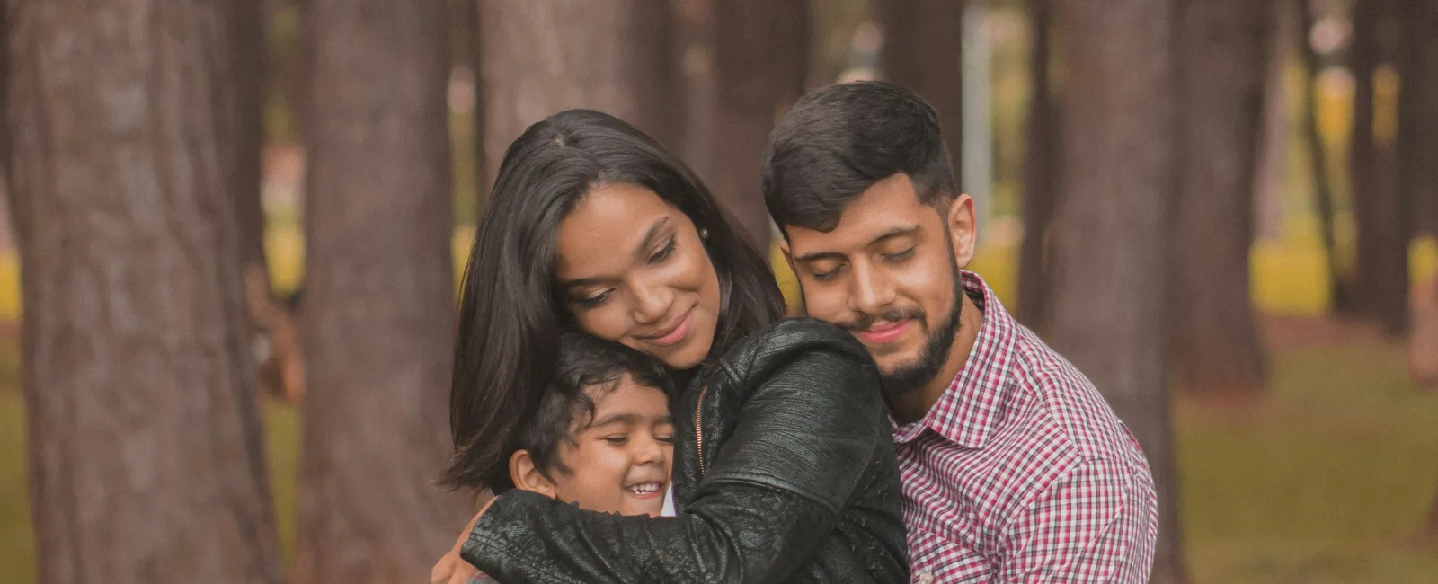 Pregnant mother with husband and child sharing a loving family hug outdoors in a wooded park area.