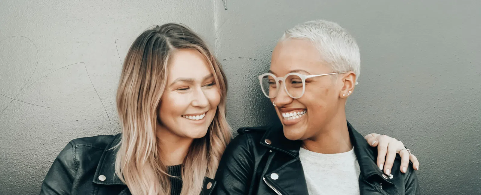 Two women in leather jackets smiling and embracing each other against a gray wall background.
