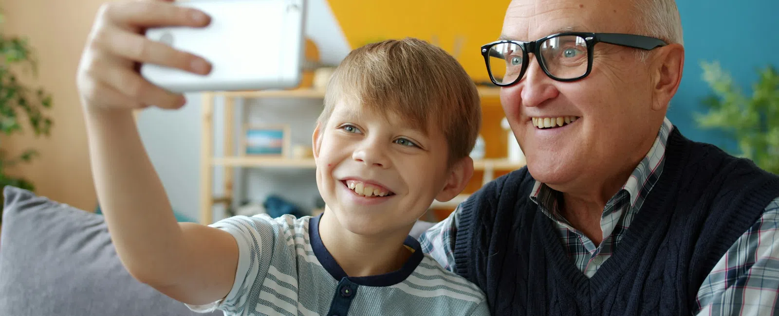 Boy and elderly man smiling together while taking a selfie with a smartphone indoors.