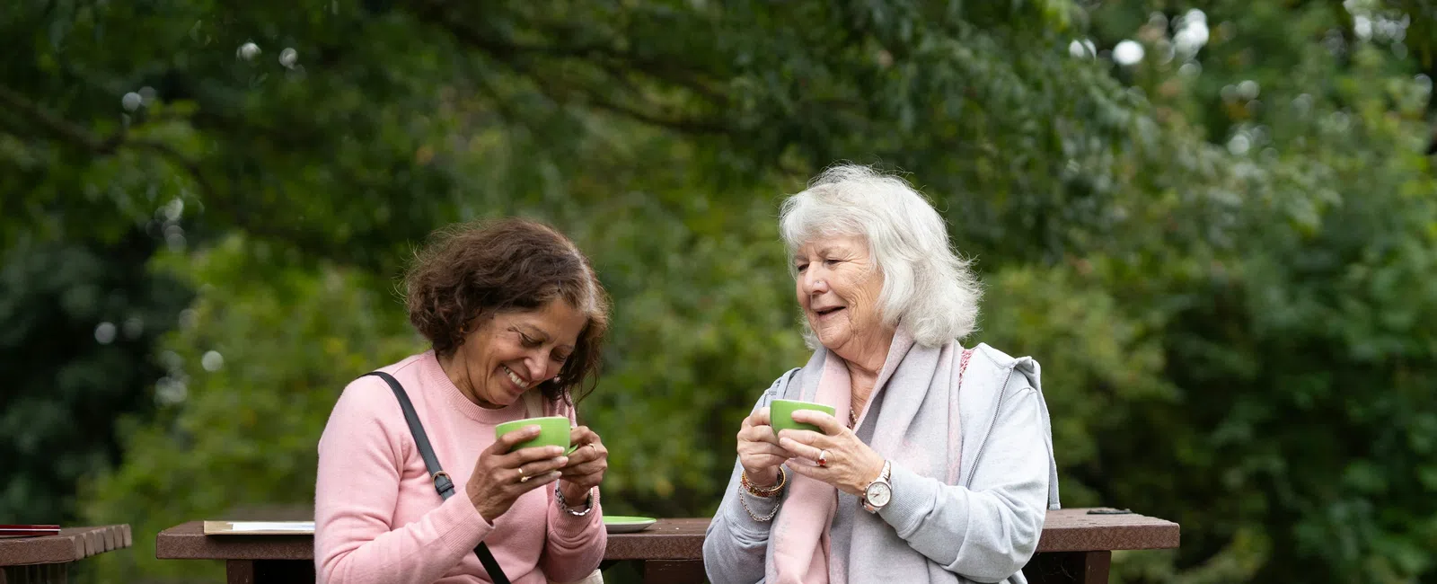 Two women enjoying tea and conversation while sitting on a wooden bench in a green park.