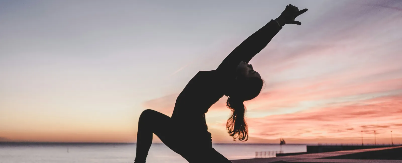 Silhouette of a woman practicing yoga in a deep stretch pose at sunset by the ocean on a mat.