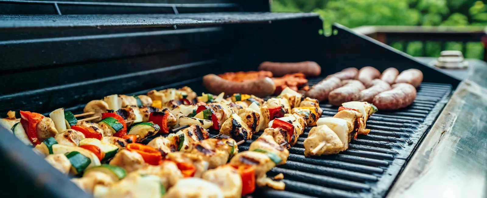 Grilled chicken and vegetable skewers with sausages cooking on an outdoor gas barbecue grill.