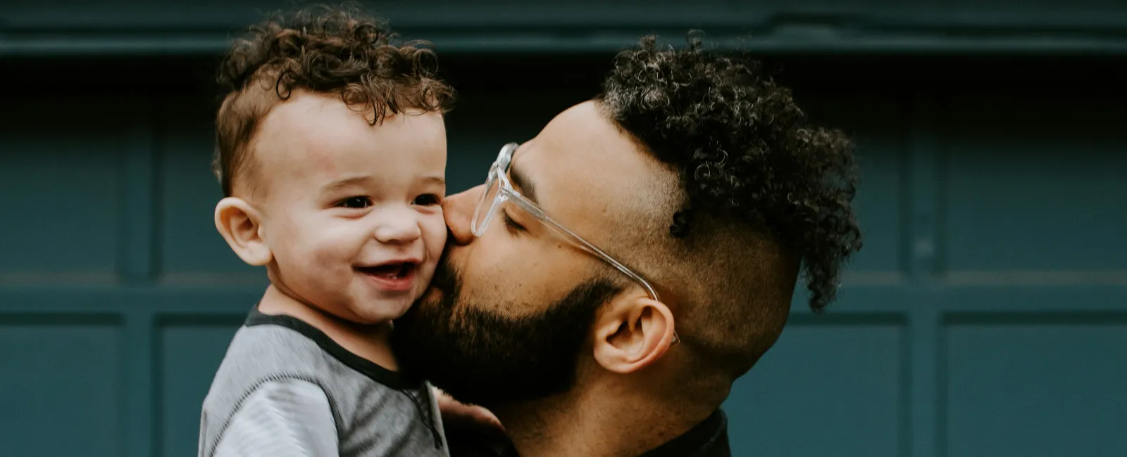 Father wearing glasses kisses smiling toddler with curly hair in front of blue garage door.