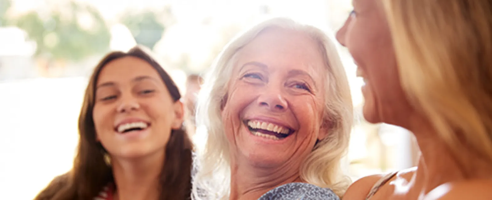 Three women of different ages happily smiling and conversing in a bright, casual setting.
