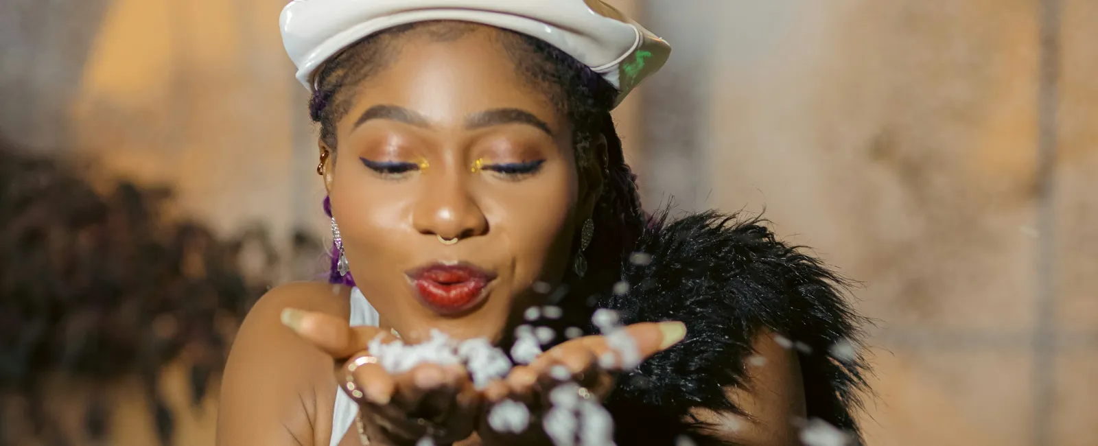Woman in white dress and beret blowing confetti with blurred indoor background and warm lighting.