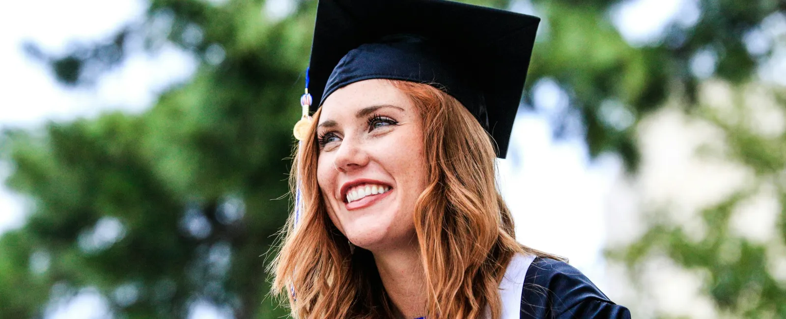 Smiling female graduate in cap and gown outdoors with blurred green trees in background