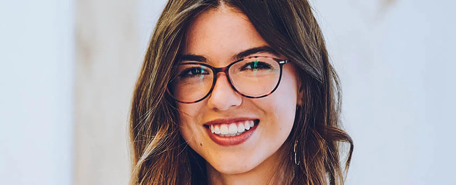 A smiling woman with long brown hair, wearing glasses and a white blouse in a bright indoor setting.