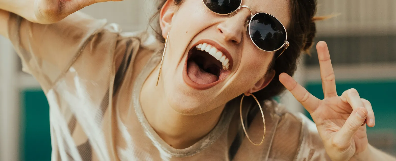 Young woman with sunglasses smiling widely and making peace signs with both hands outdoors