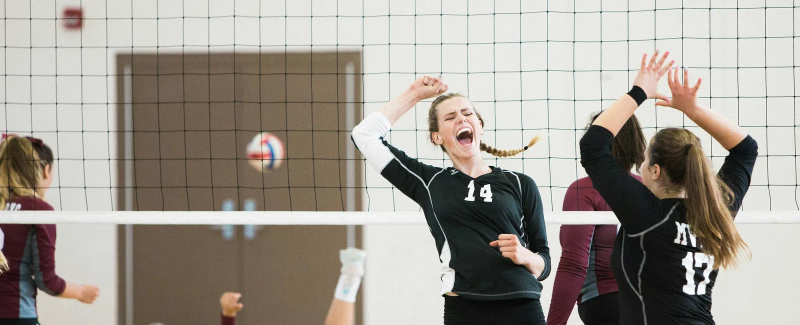 Women volleyball players in black and maroon uniforms playing an indoor game with one player celebrating near the net.