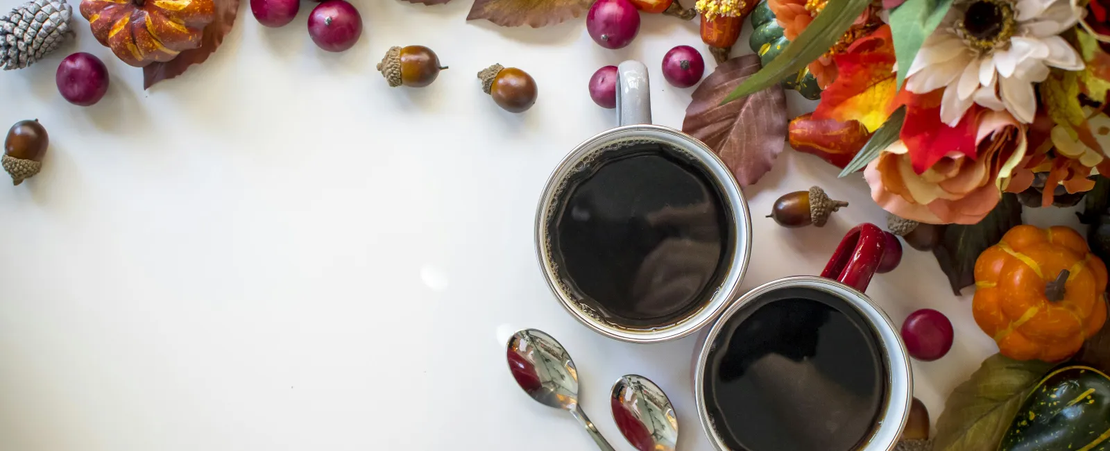 Two cups of coffee surrounded by autumn leaves, pumpkins, pinecones, acorns, and decorative flowers on white.