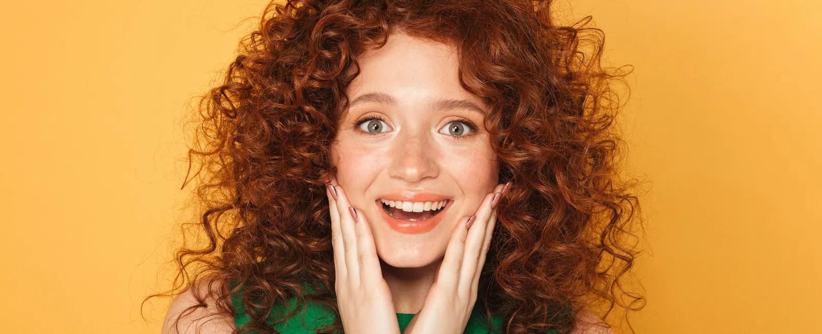 Surprised redhead woman with curly hair wearing green top against yellow background