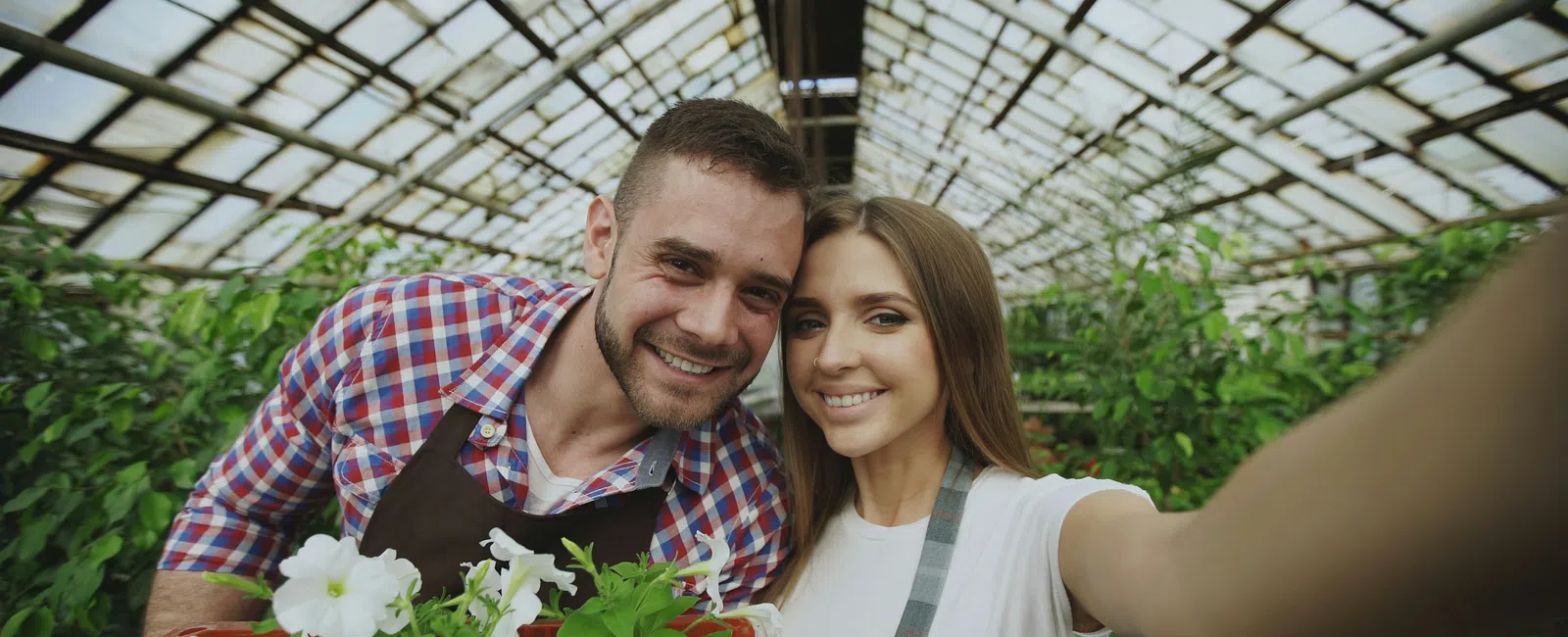 Smiling couple taking a selfie in greenhouse with white flowers and plants in red crate, wearing aprons