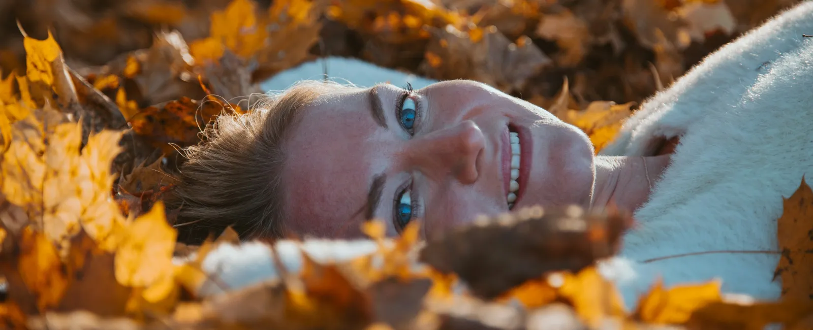 Smiling woman with blue eyes lying on ground covered in yellow autumn leaves wearing a white sweater.