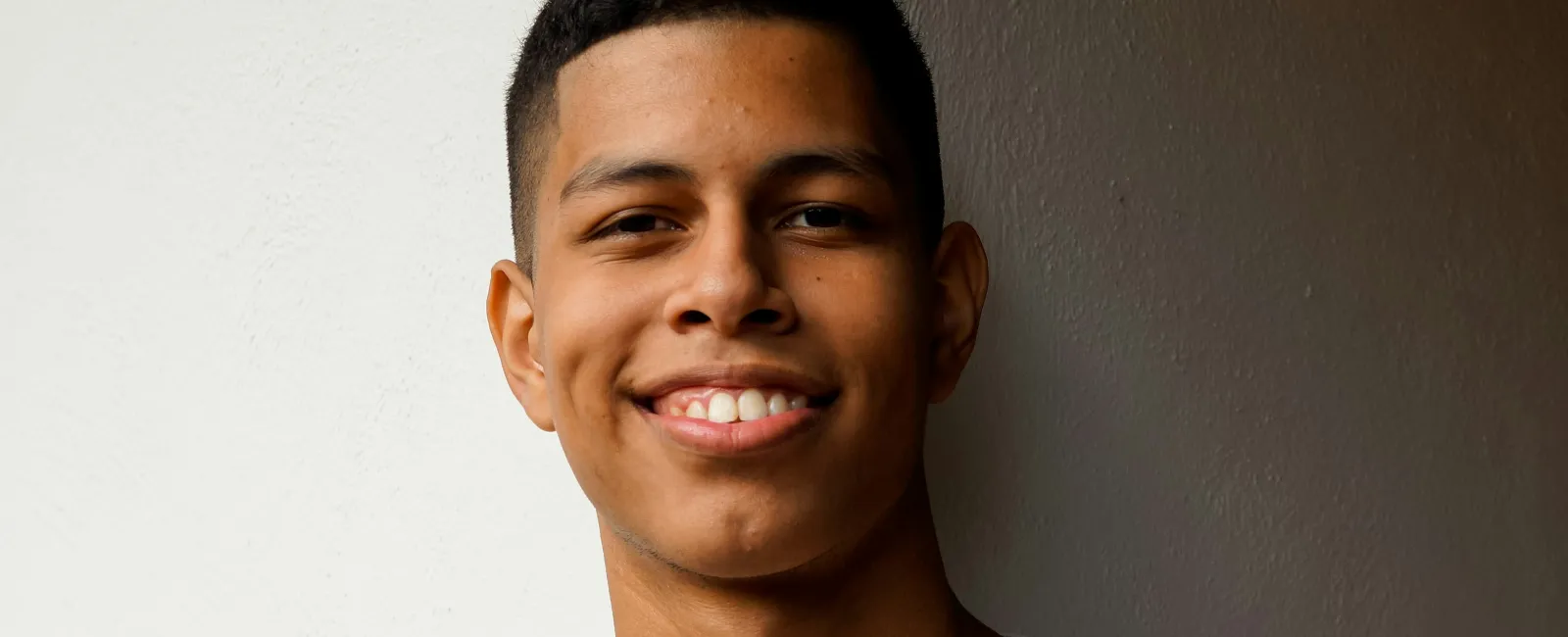 Smiling young man in a light beige t-shirt standing against a wall with white and dark brown sections.
