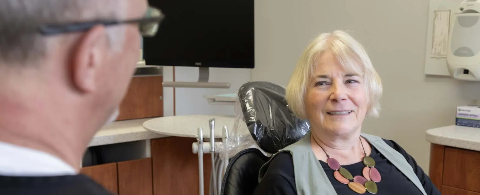 Senior woman smiling while sitting in dental chair talking to dentist in modern clinic.