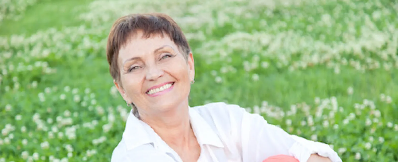 Smiling mature woman sitting cross-legged in a green field of white flowers wearing a white shirt and coral pants.
