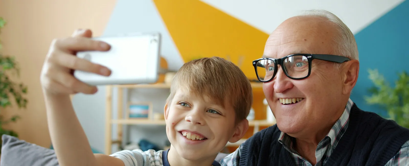 Smiling grandfather and grandson taking a selfie indoors with colorful geometric wall in the background