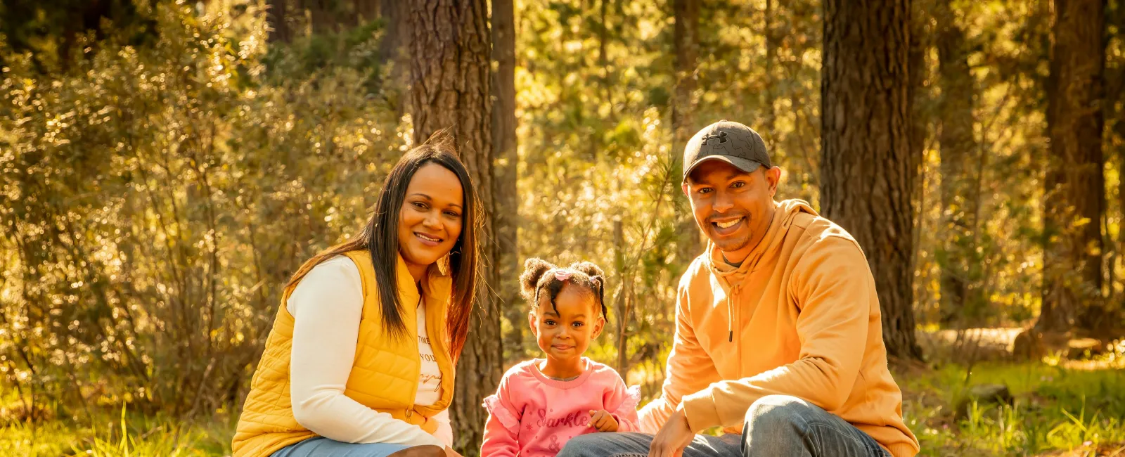 Smiling family of three sitting on a log in a sunlit forest during autumn outdoor outing