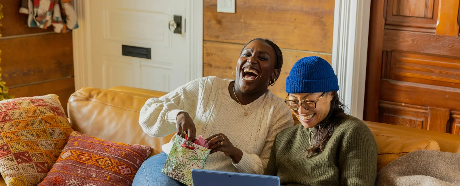 Two friends laughing on a couch while using a laptop and opening a gift bag in a cozy living room.