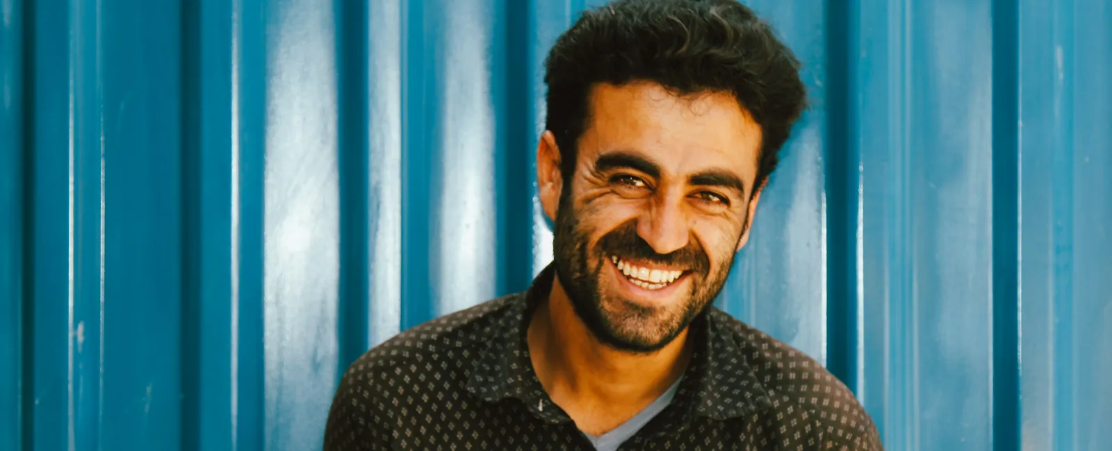 Smiling man with dark hair and beard standing in front of blue corrugated metal wall wearing a patterned shirt.
